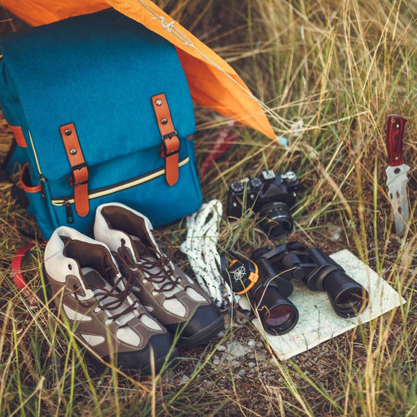 Blue backpack, hiking boots, binoculars, and a map on grass near a tent.