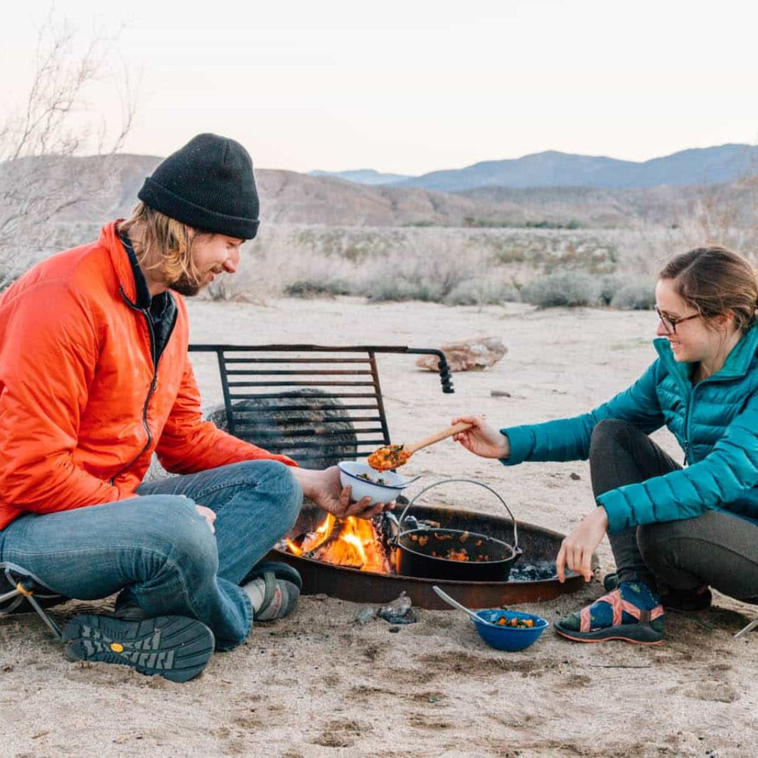 Two people cooking outdoors by a campfire with mountains in the background