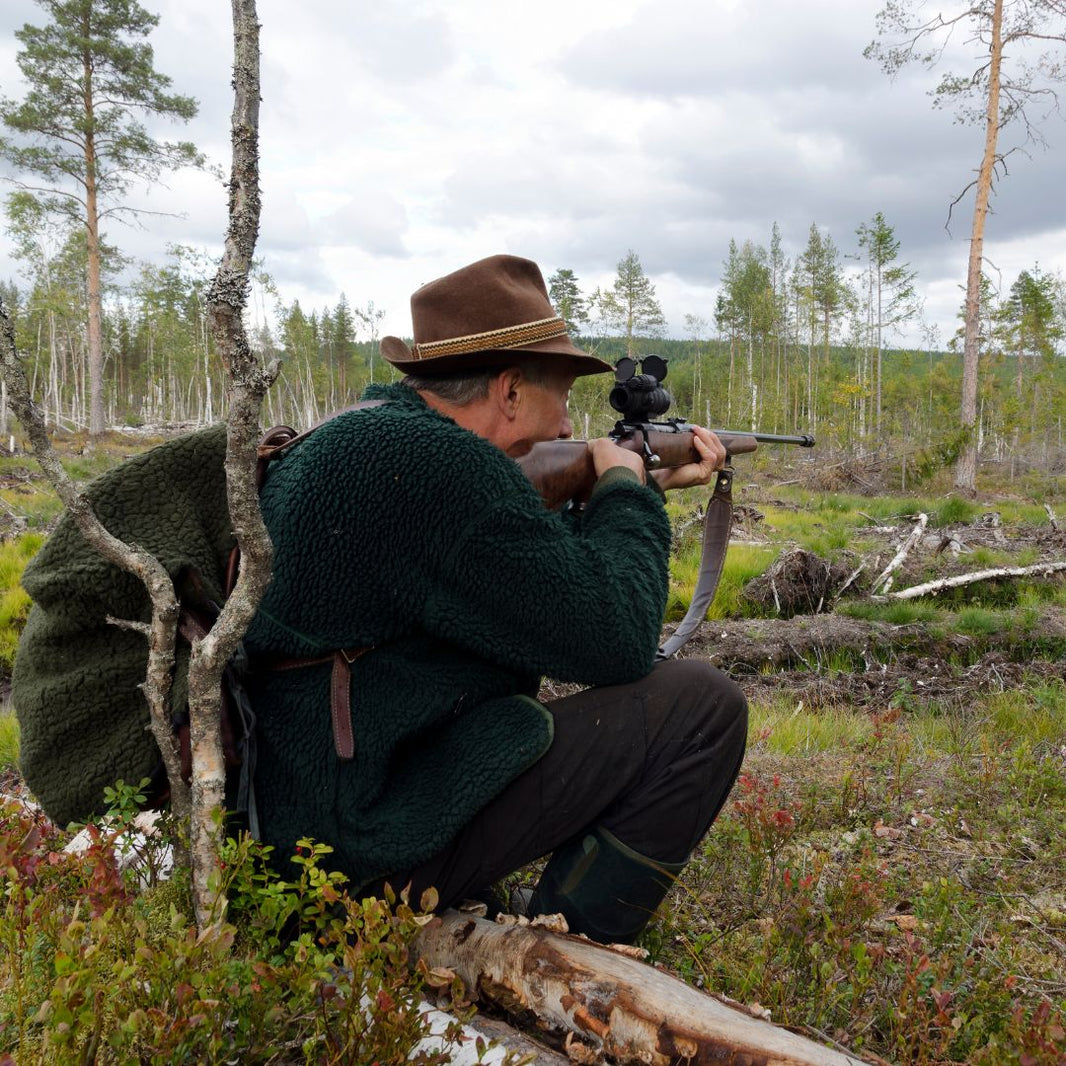 Person in a forest setting with a rifle, wearing a green jacket and brown hat.