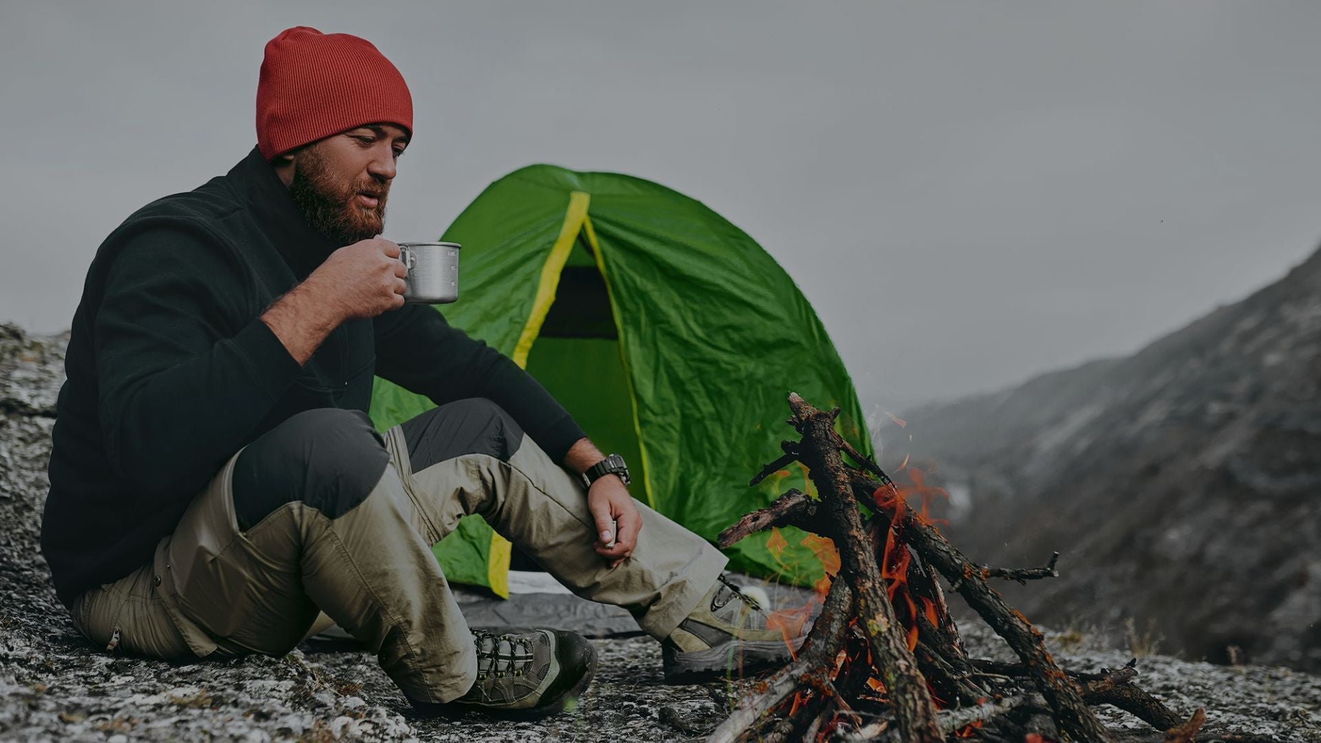 Man camping in a mountainous area, sitting by a campfire with a green tent in the background.