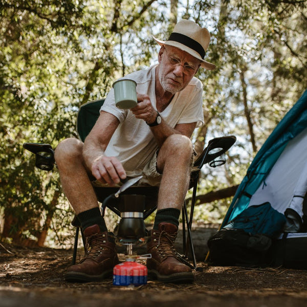 Man sitting outdoors with camping gear, holding a cup.