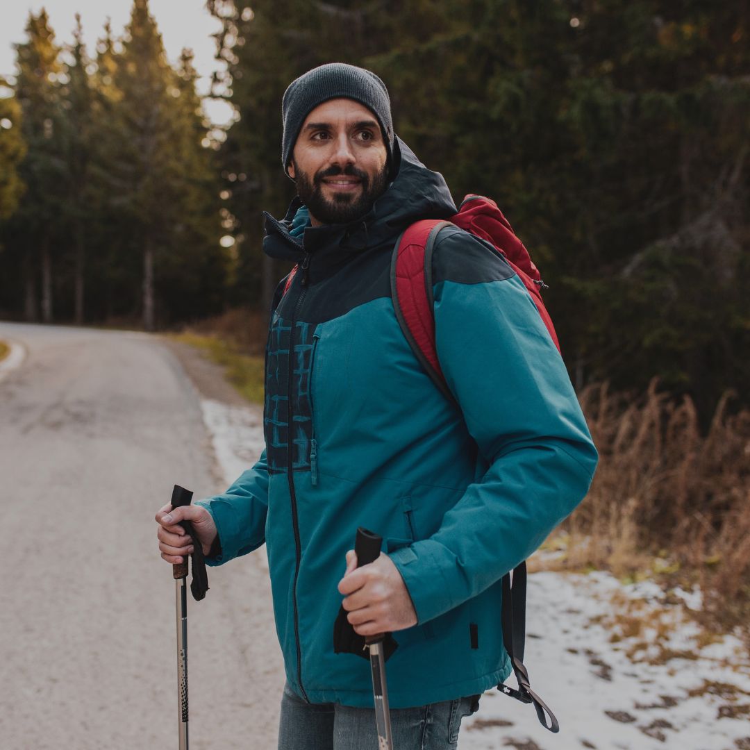 Man in a blue jacket with a red backpack standing on a snowy path with trees in the background