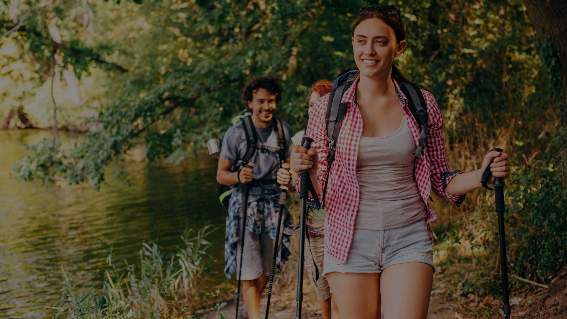 Two hikers with backpacks and walking sticks walking along a path by a body of water.